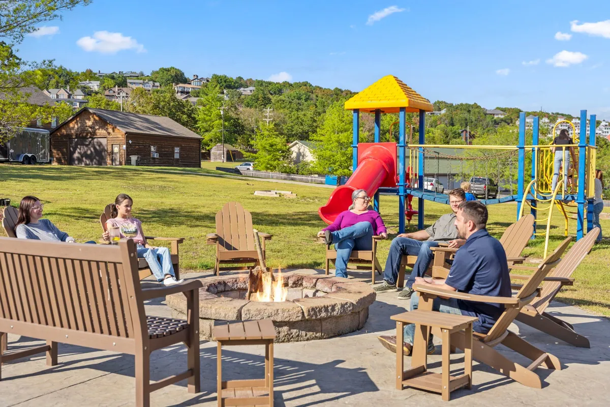 A fire pit gathering area with comfortable seating, a playground visible in the background, and people enjoying the warmth of the fire under the evening sky. Children play nearby while adults relax by the fire, capturing a sense of togetherness and relaxation.