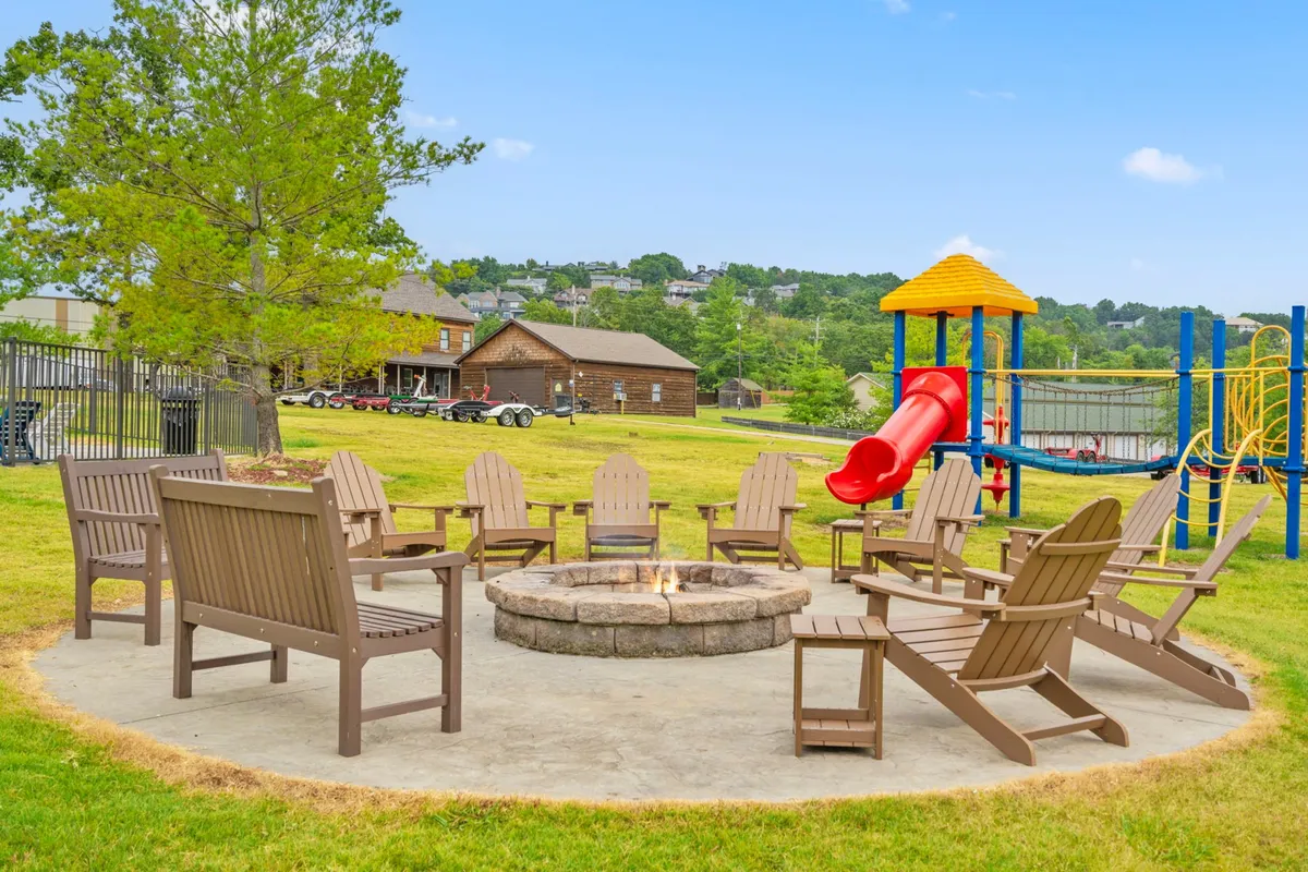 A cozy fire pit surrounded by Adirondack chairs sits near a playground slide. The area is filled with the warm glow of a crackling fire as families gather, children play, and the starry night begins to fall.