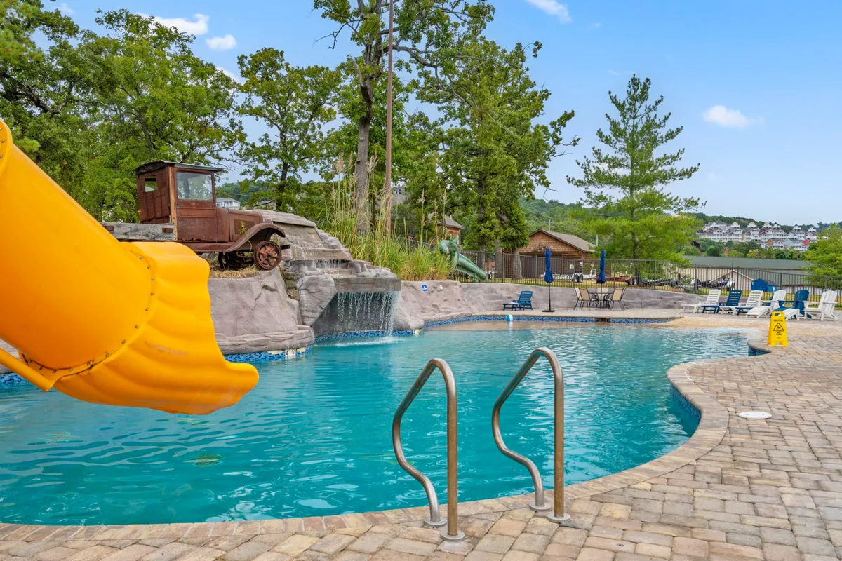 A vibrant outdoor pool scene featuring a sparkling swimming pool with a yellow slide, a waterfall feature, a rustic old truck, and plenty of lounge chairs. The sun is shining, and the area is set up for relaxation and fun, with families and children visible enjoying the water and the outdoor space.