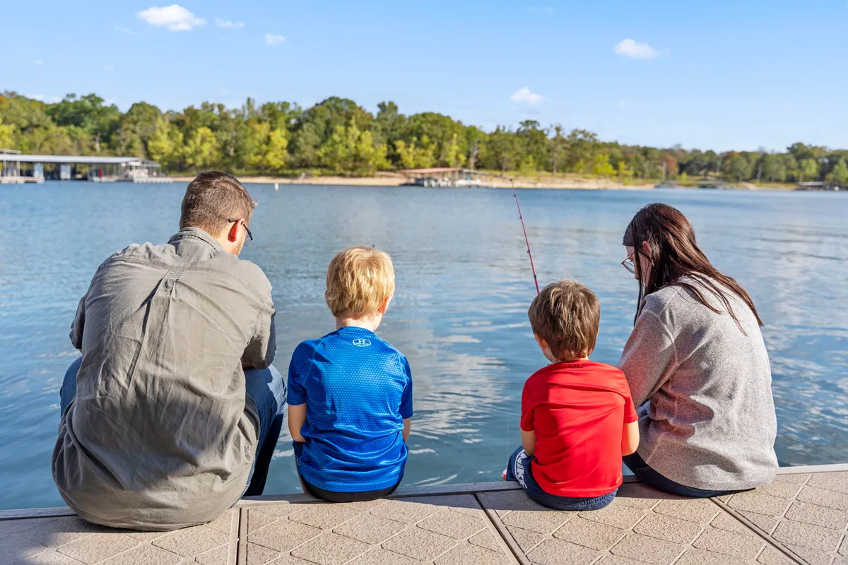 A family sits on the shore by the calm lake, with children fishing and beautiful reflections on the water. The scene conveys joyful family bonding and a peaceful, welcoming environment for both people and pets.