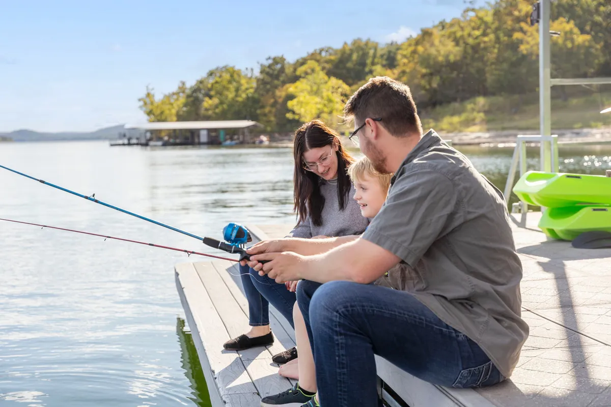 A cheerful family stands on a dock, casting their fishing rods into the sparkling lake. The scene is filled with laughter and togetherness as one child proudly displays a newly caught fish, while lush greenery surrounds the peaceful water.
