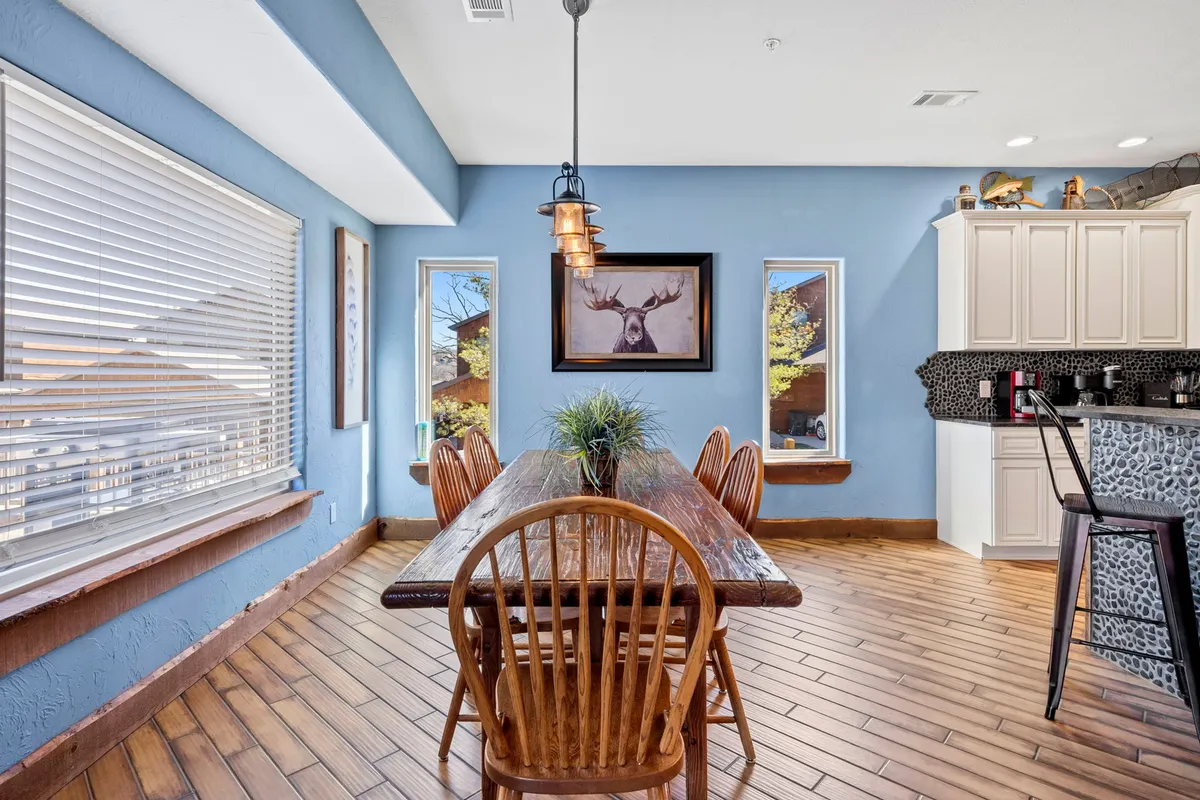 A warmly lit dining area featuring a rustic wooden dining table set beneath large windows, with an antler-themed painting adding character. The table is ready for family-style meals or celebrations, creating a welcoming atmosphere.