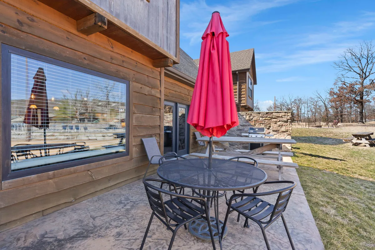 An inviting outdoor patio scene featuring a red umbrella, an outdoor dining table set for a meal, and a charming stone wall in the background. The area is surrounded by greenery, creating a cozy atmosphere for al fresco dining and relaxing.