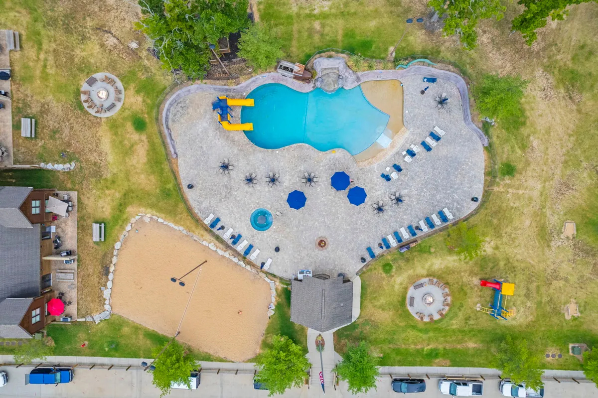 A vibrant pool area featuring water slides and a sandy volleyball court is framed by lush greenery and comfortable sitting areas. Families and friends are seen swimming, sunbathing, and playing volleyball, creating a lively and inviting atmosphere.