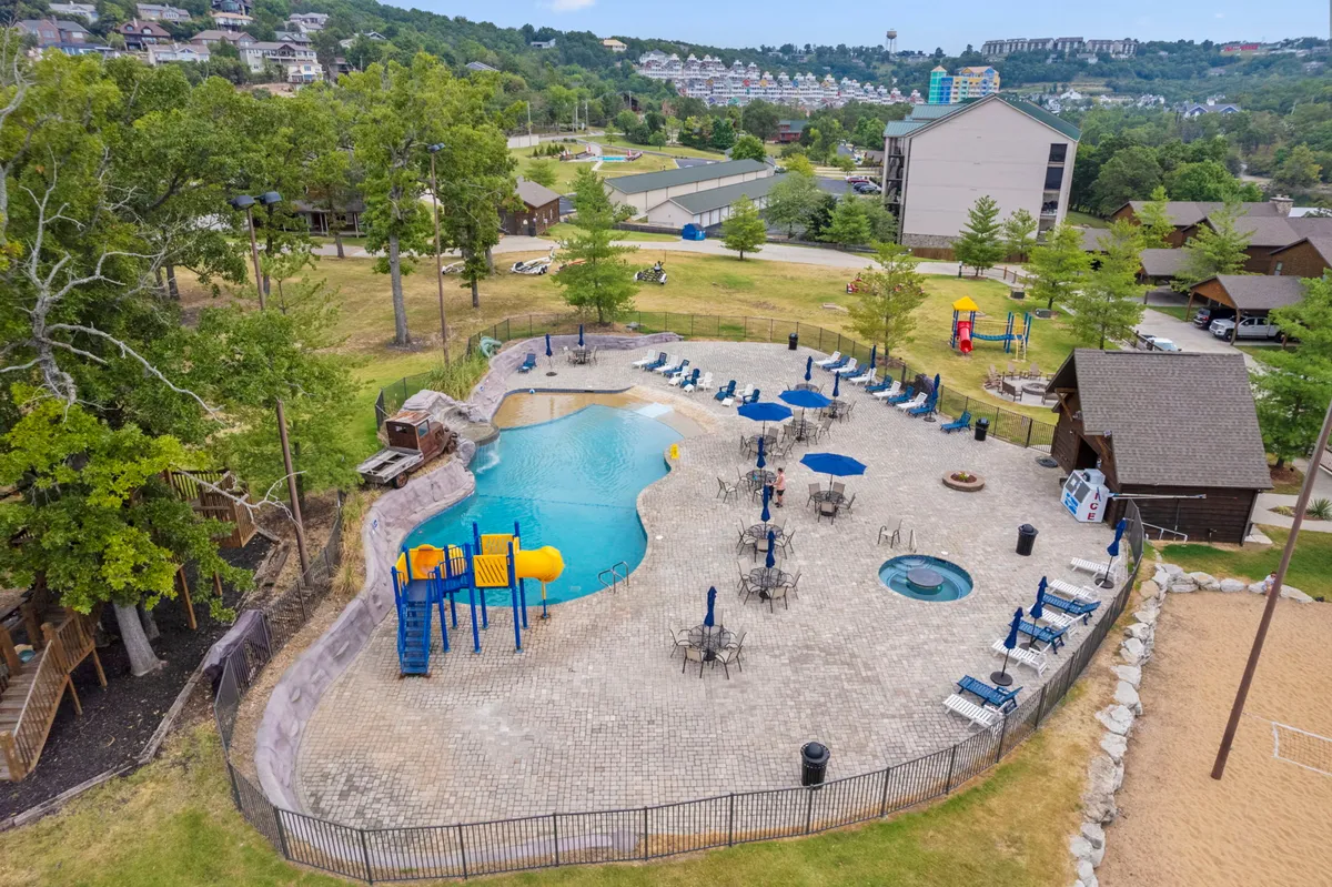 A sweeping view of the resort's social hub, showcasing a swimming pool, adjacent playground, hot tub, seating areas, and a sand volleyball court. Families and children are visible enjoying the amenities, embodying the vibrant, recreational spirit of the area.