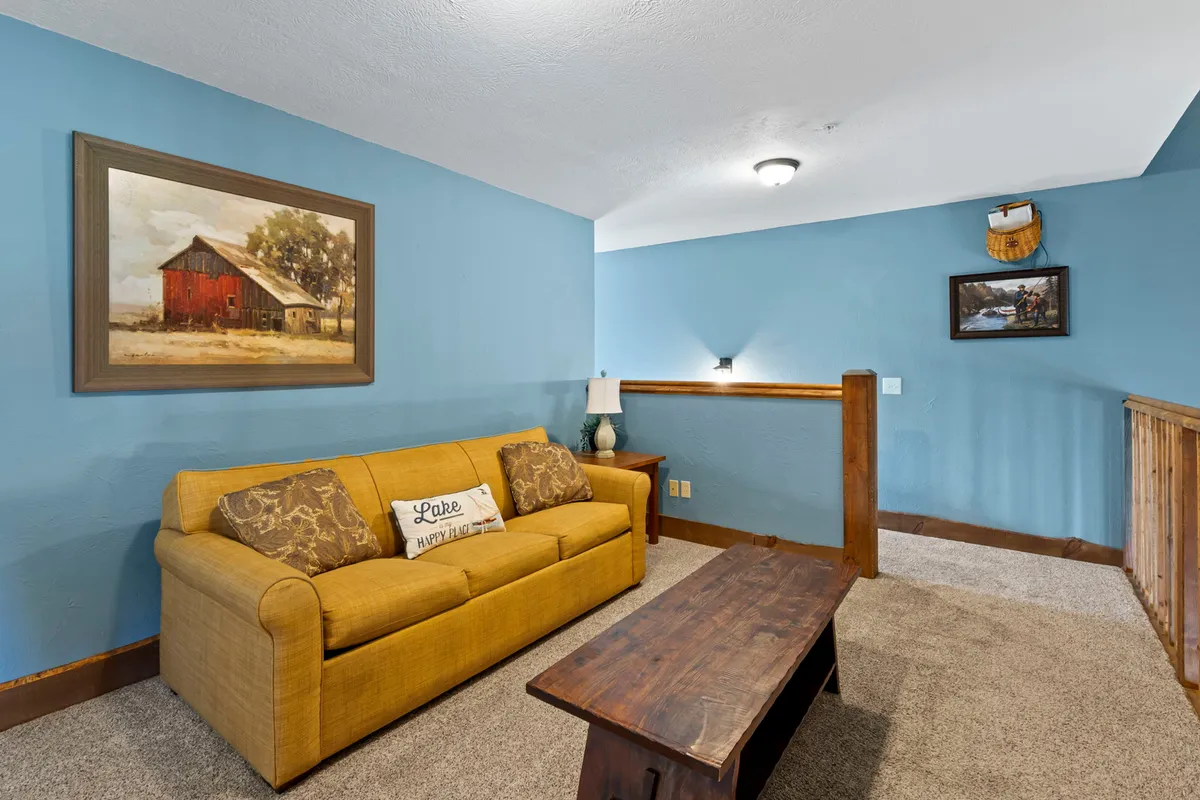 A cozy loft area with a yellow couch, wooden coffee table, and framed artwork. A lamp adds warm lighting, creating an inviting spot for reading, relaxing, or extra sleeping arrangements for guests.