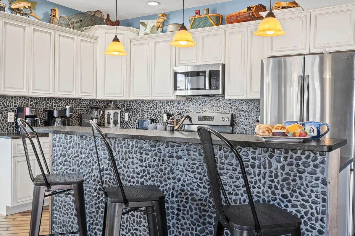 A close-up view of the kitchen's stone-patterned bar counter with hanging pendant lights, modern appliances, and a breakfast display on the counter. Decorative shelves and inviting lighting make the space feel homey and functional.