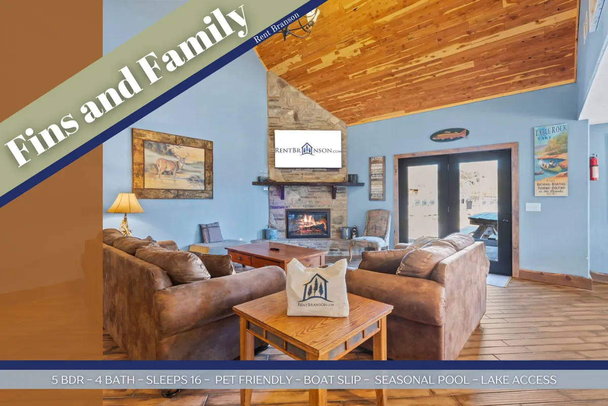A welcoming open-plan living area featuring comfortable sofas arranged around a stone fireplace, a large TV mounted above the mantle, and decorative wooden ceiling beams. Lake-themed decorations and a spacious layout create a warm, communal atmosphere ideal for group activities.