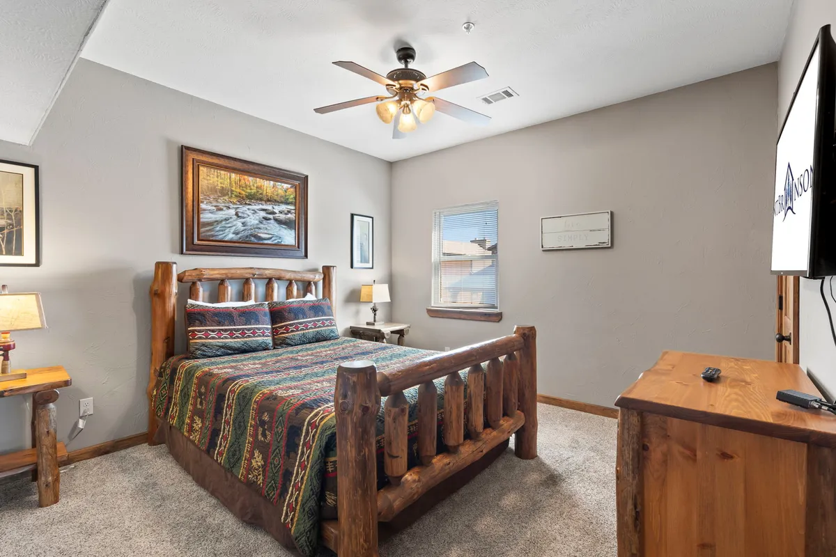 A cozy bedroom with a log bed, colorful blanket, and rustic artwork on the wall. A lamp sits on the nightstand, and a television is mounted for entertainment. Natural light streams through the window, creating a serene and inviting atmosphere.