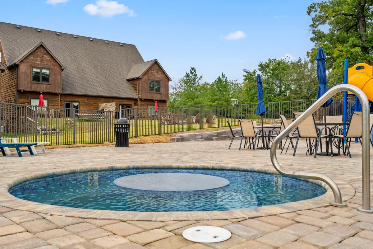A cheerful backyard scene showing a hot tub beside a pool area, surrounded by patio seating and a playground slide nearby. The space is set up for family gatherings, outdoor dining, and relaxation, with children playing in the background and a welcoming vibe.