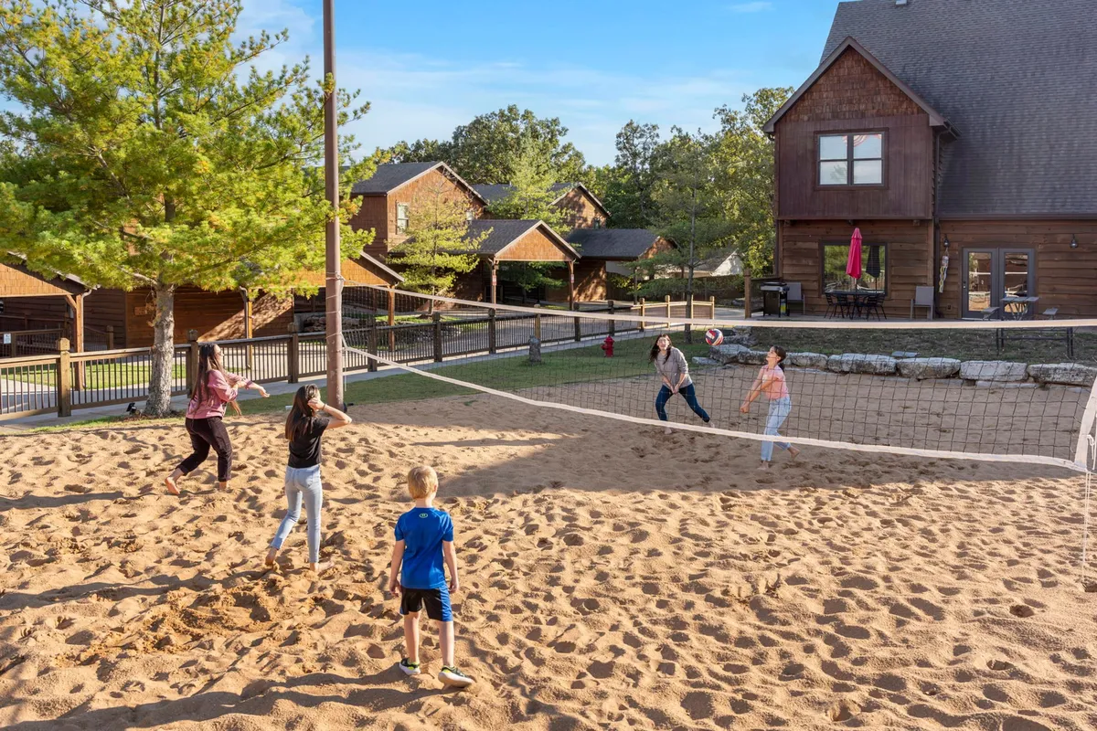 An energetic sand volleyball court bordered by wooden cabins and lush trees, with children actively playing volleyball. The scene is lively and filled with laughter, capturing outdoor community and friendly competition.