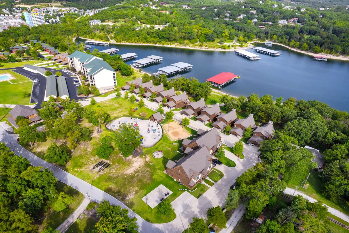 A vibrant exterior scene featuring the lake, boathouses, a playground, residential cabins, and a swimming pool. The area is alive with opportunities for boating, picnicking, swimming, and nature walks, all set against a backdrop of water and greenery.